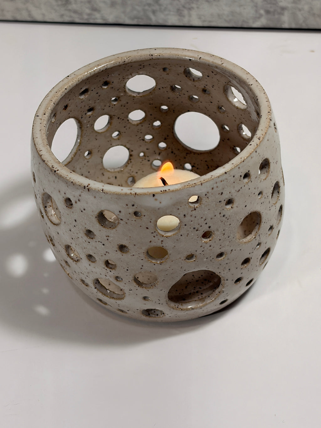 A speckled ceramic candle holder with a lit tea light candle inside, set on a white surface with a soft shadow.
