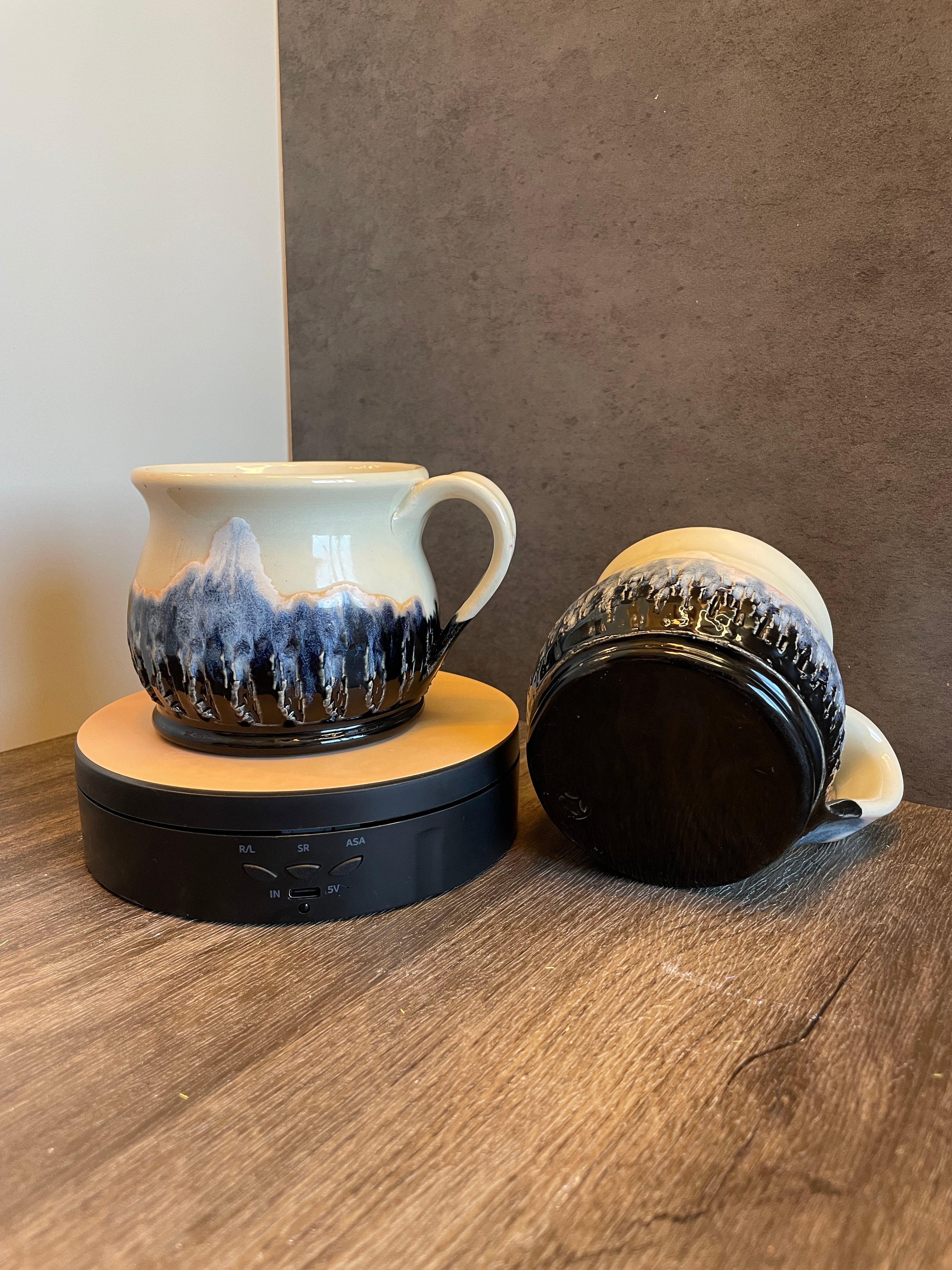 A ceramic mug with a mountain landscape design in blue, black, and cream, shown on a wooden surface with its black lid beside it.
