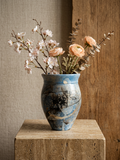 A blue and gray marbled ceramic vase filled with dried flowers and greenery, displayed on a stone pedestal against a textured beige and wood background.
