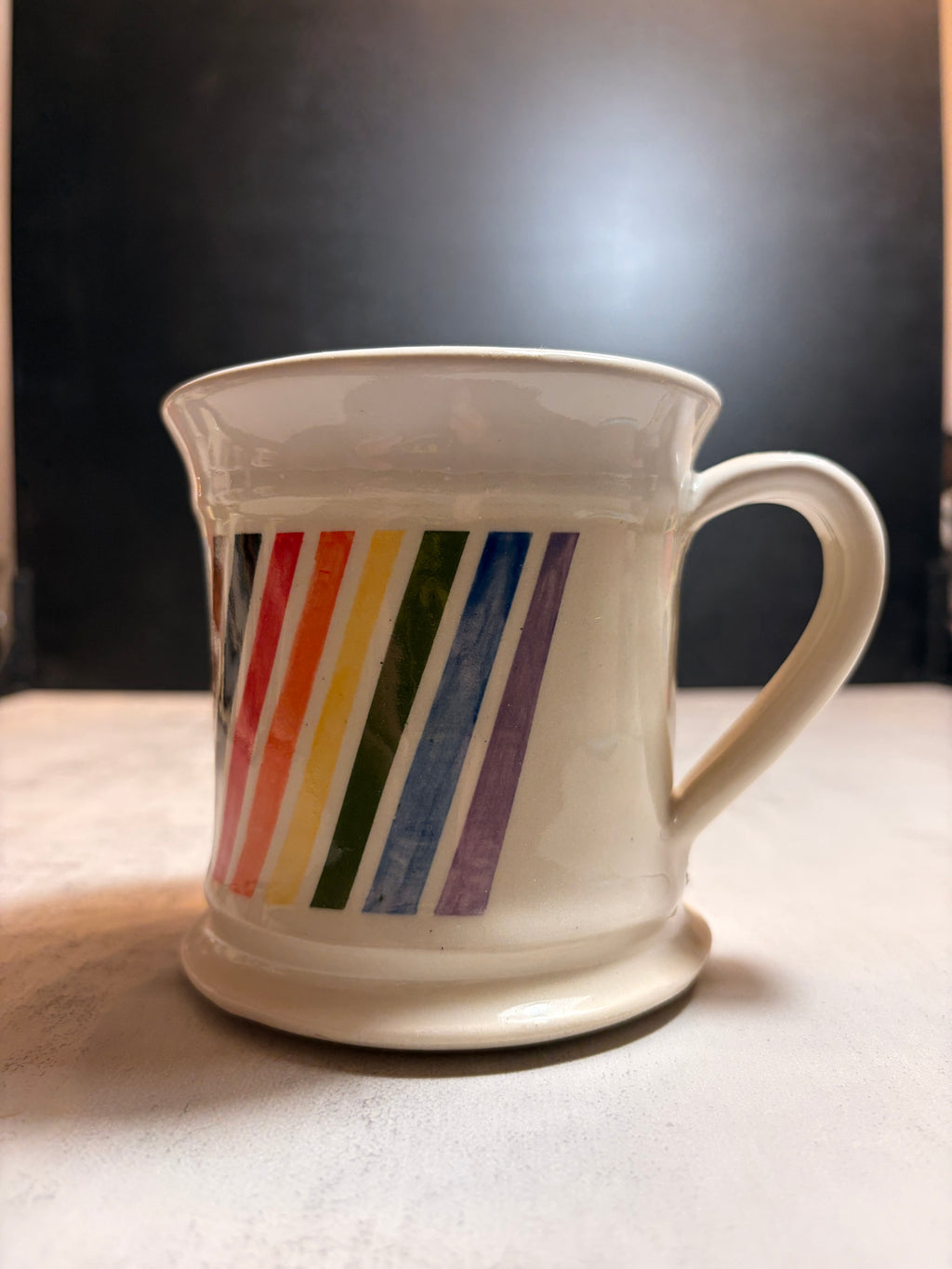 A white ceramic mug with a handle and base, decorated with diagonal rainbow stripes in red, orange, yellow, green, blue, and purple, set against a dark background.