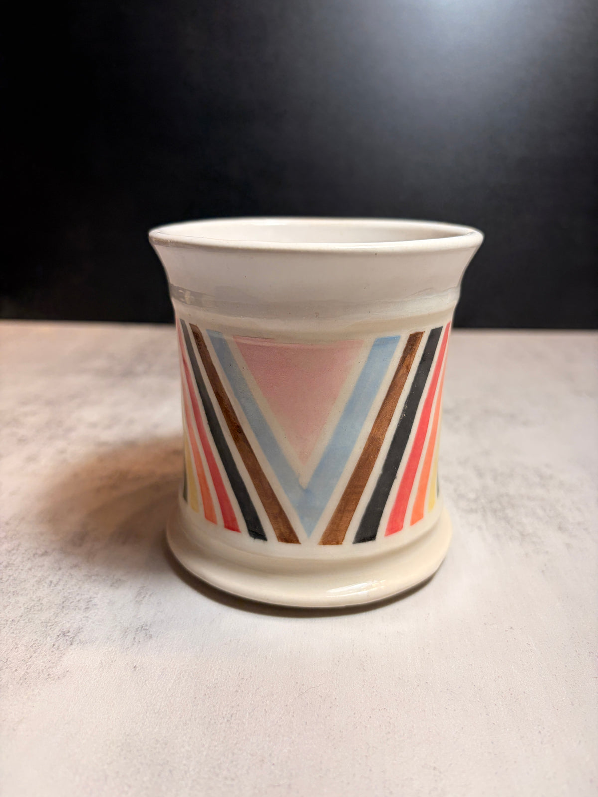 A small ceramic cup with a white base and a colorful geometric pattern featuring pink, blue, orange, brown, and black stripes, set on a light gray surface against a dark background.