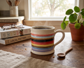 A ceramic coffee mug with colorful horizontal stripes sits on a wooden table in a cozy room setting, with a potted plant, glasses, and books nearby.