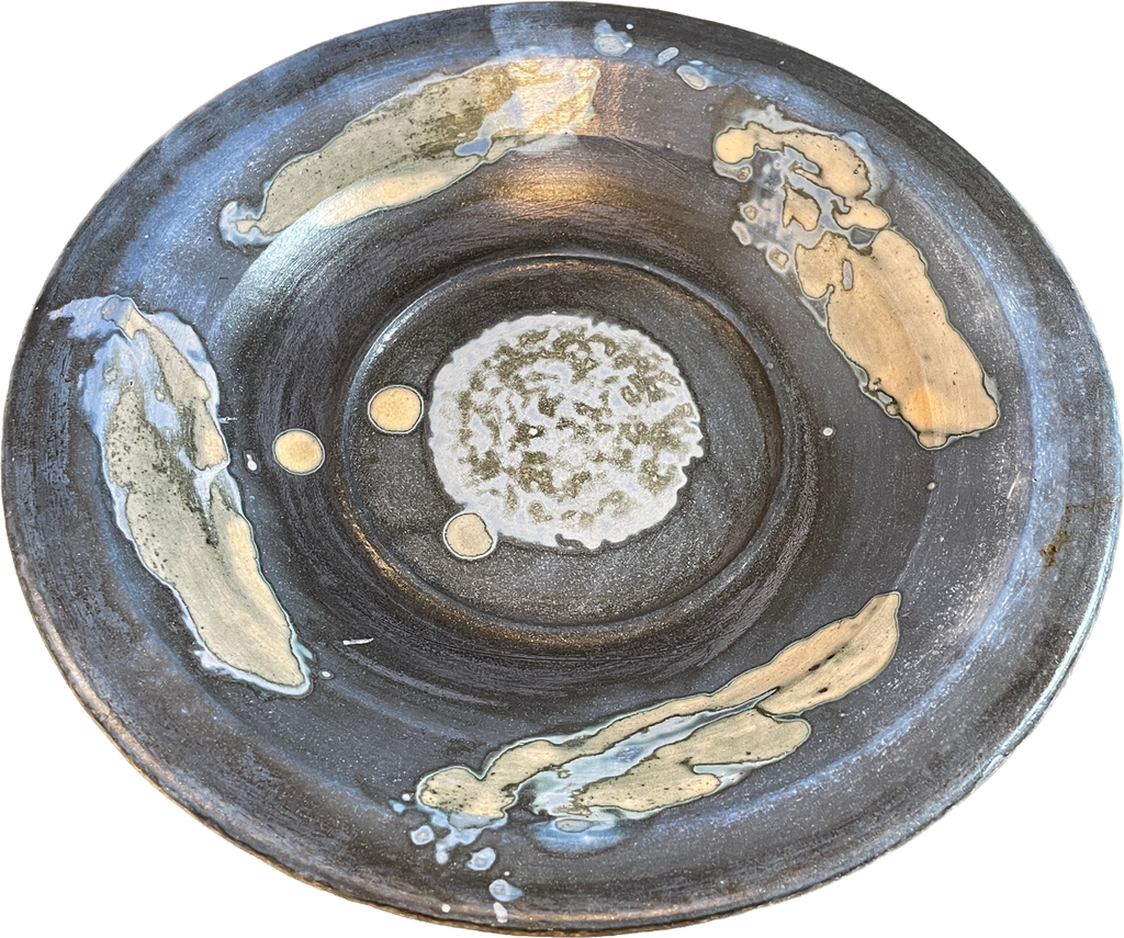 A round handmade ceramic plate with a dark charcoal gray glaze, featuring abstract hand-painted gold and beige brushstrokes and a textured white and green center.